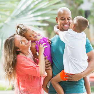 A man and a woman with two small children embrace in front of palm trees.